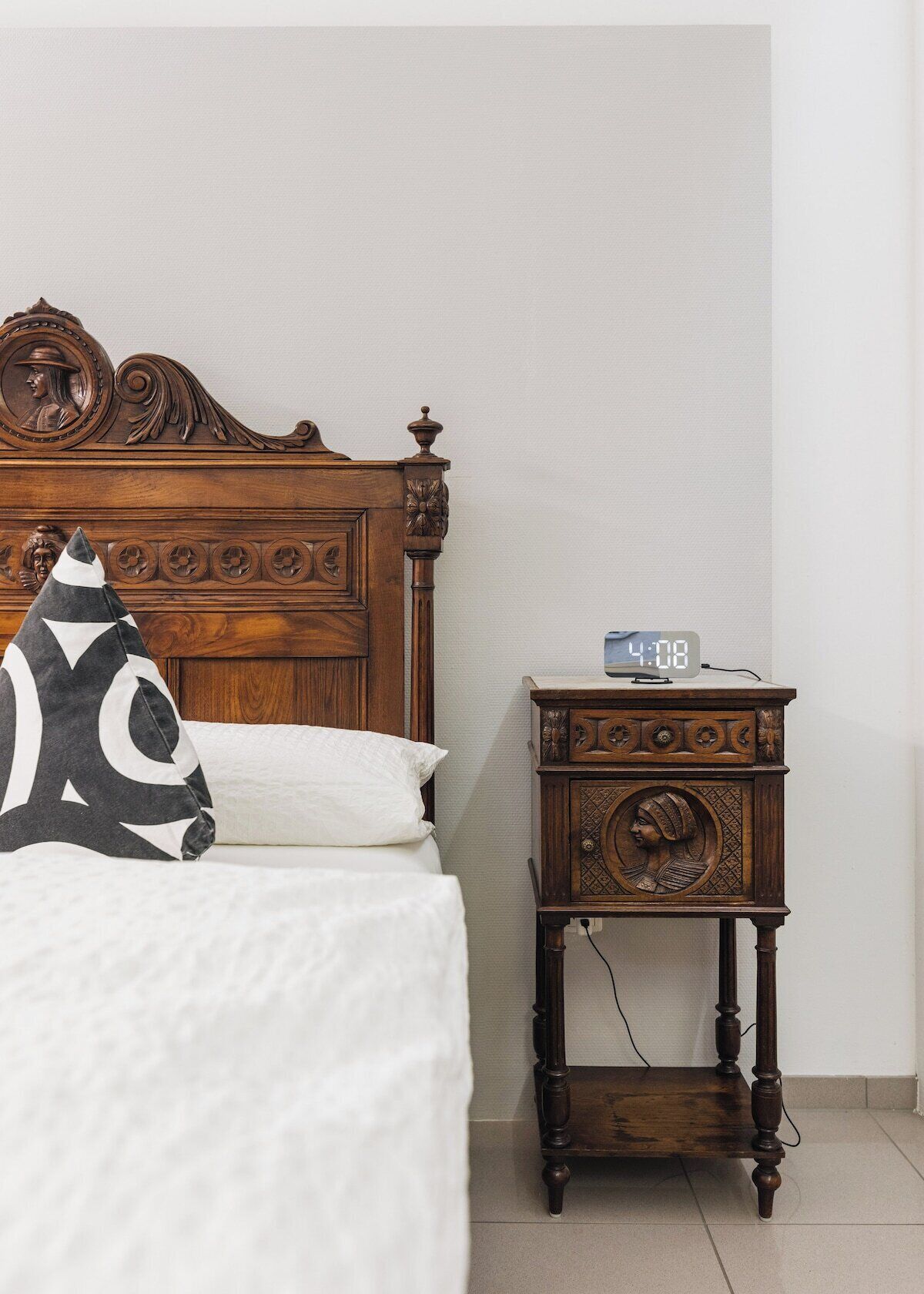 A wooden bed with a carved headboard, a bedside table with a clock, and a pillow with a geometric pattern.