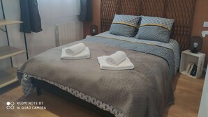En pænt redt seng med mønstret sengetøj og to rullede håndklæder, et natbord med en lampe, og et vindue med gardiner.