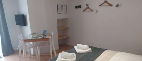 A neatly arranged bedroom with a bed, a desk, and a chair. There are towels on the bed and a small shelf unit against the wall.