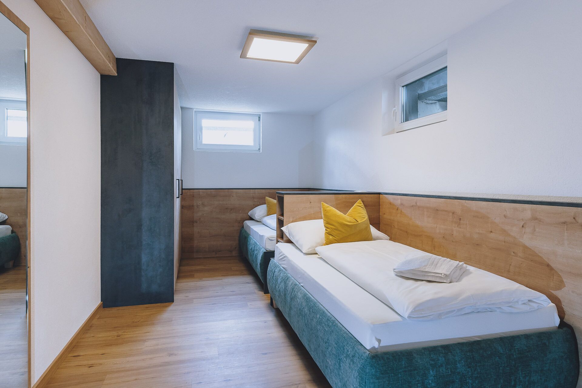 Habitación de hotel con cabecera de madera, una cama individual con ropa blanca y almohadas amarillas, una ventana pequeña y suelo de madera.