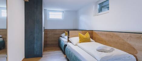 Habitación de hotel con cabecera de madera, una cama individual con ropa blanca y almohadas amarillas, una ventana pequeña y suelo de madera.