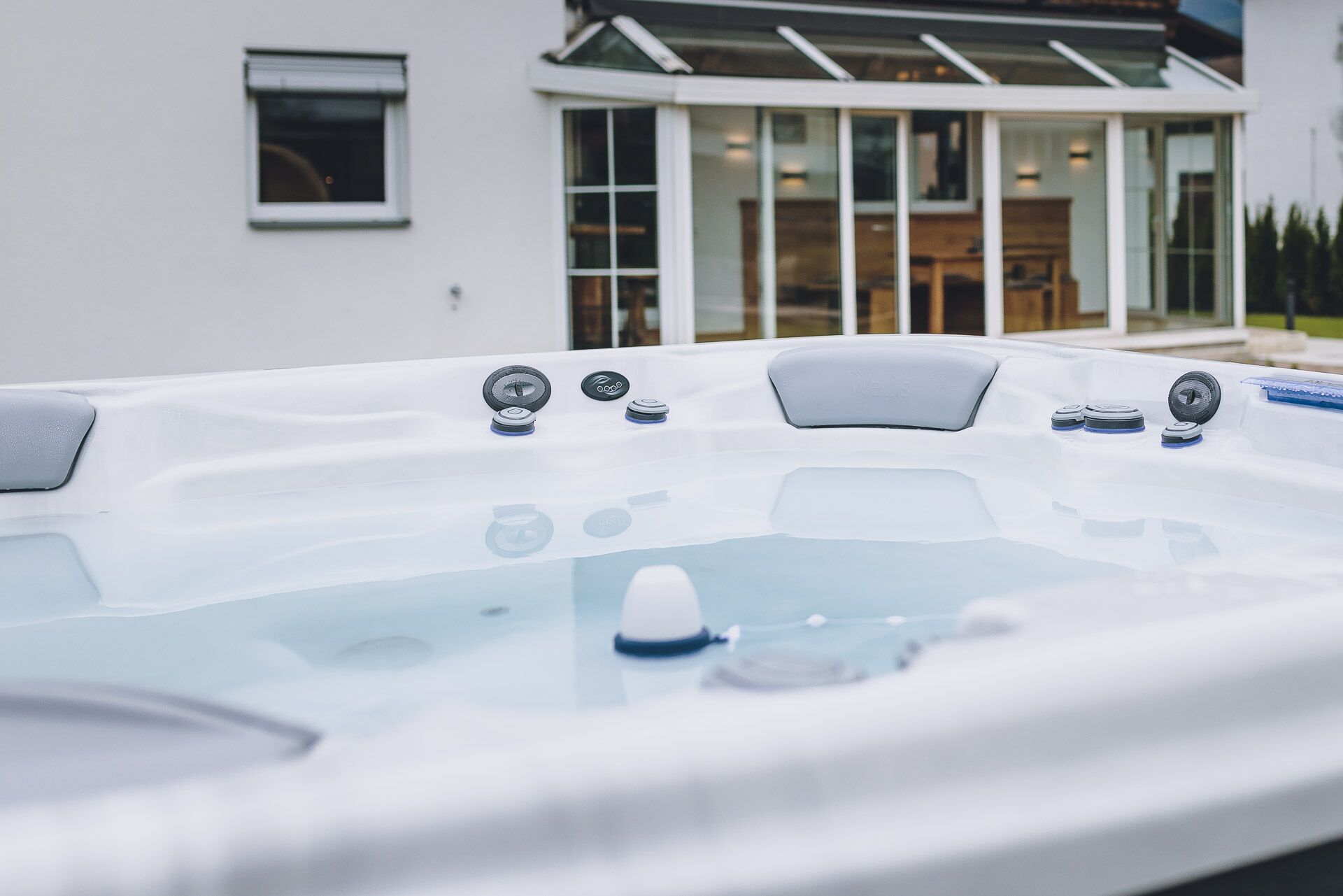 Un bain à remous doté de commandes et de jets, installé devant une maison moderne avec de grandes fenêtres et une porte en verre.