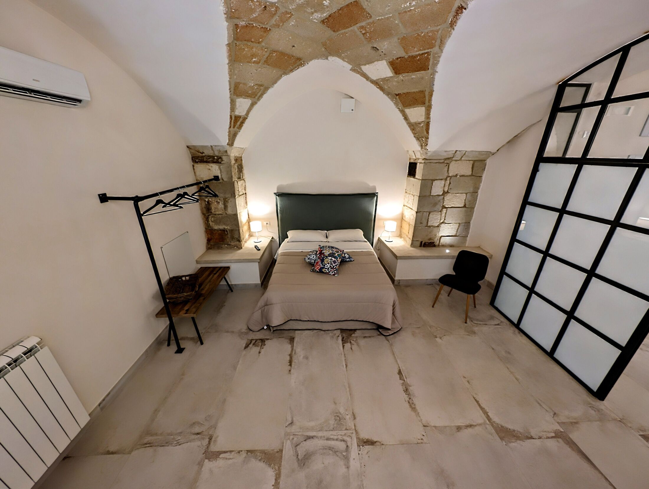 Quarto com teto de pedra, cama com cabeceira verde, luminárias de cabeceira, uma cadeira e um radiador.