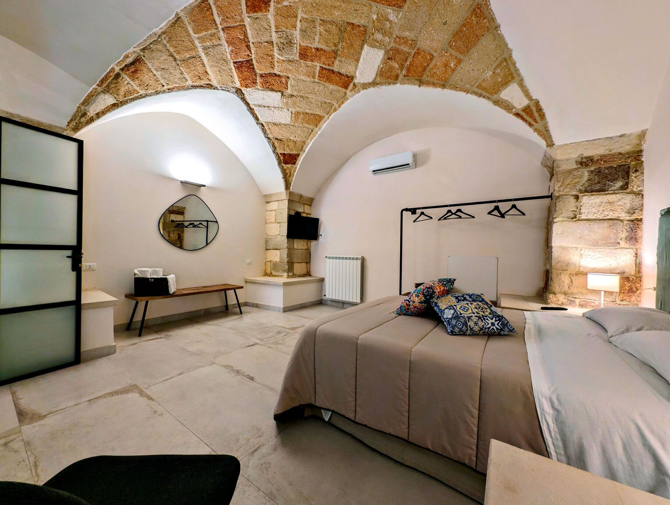 Ein Schlafzimmer mit einer Decke aus Steinbögen, einem Bett mit gemusterten Kissen, einem Nachttisch, einem Spiegel und einer Hängelampe.