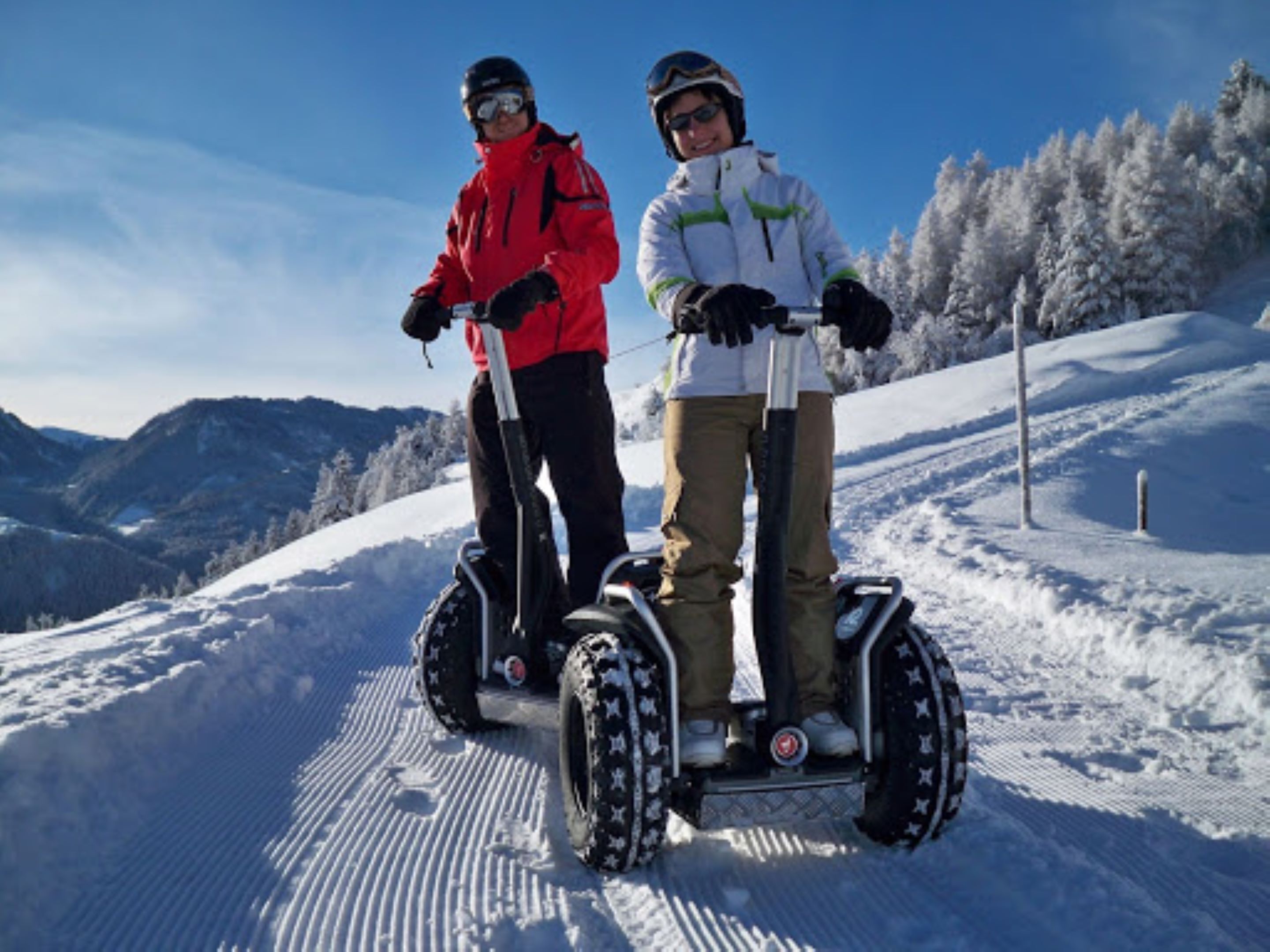 Two people on Segways on a snowy slope with mountains in the background.