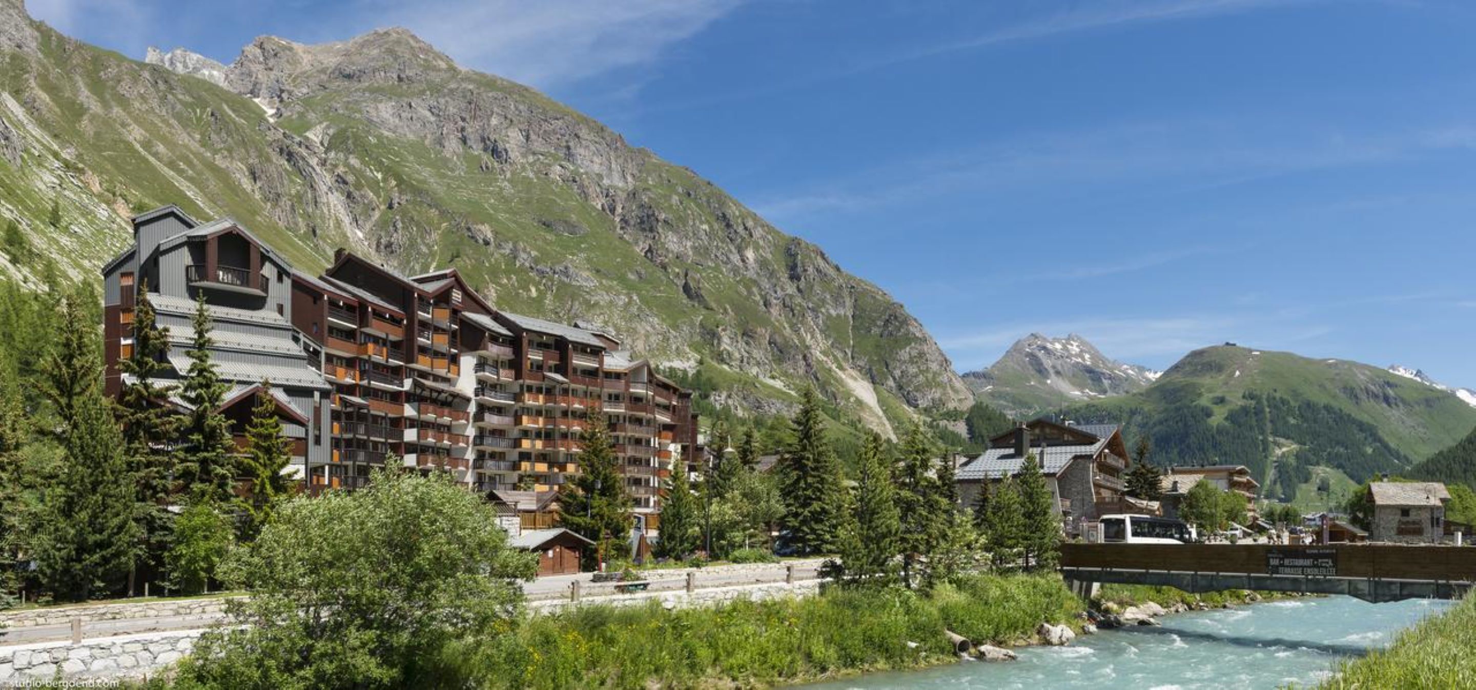 Un hotel a più piani con balconi, un fiume e un paesaggio montuoso sullo sfondo.