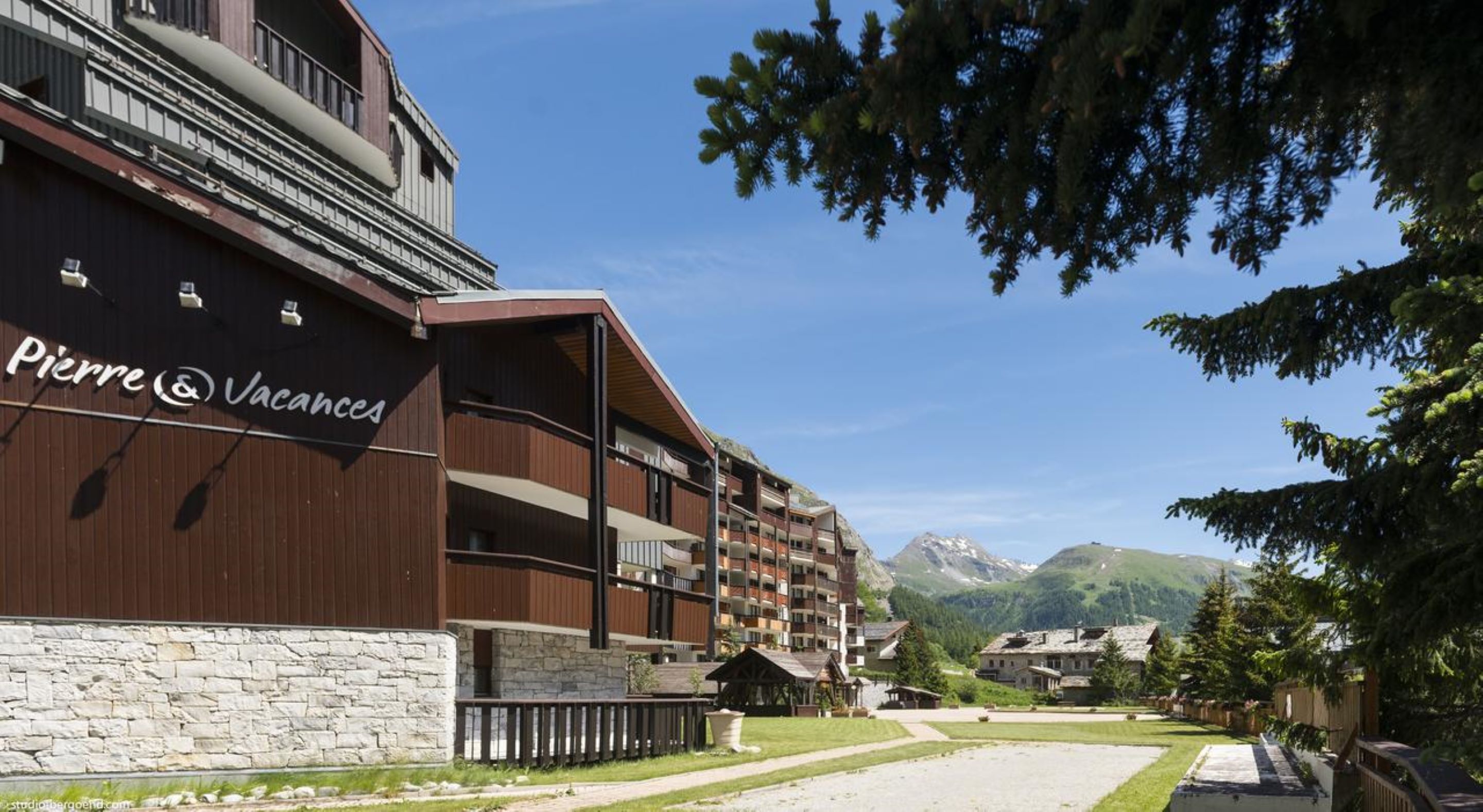 Un hotel moderno con varios balcones, fachada de piedra y vistas a las montañas al fondo.