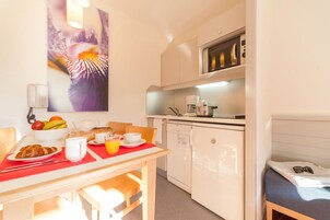 Cocina compacta con microondas, cafetera y refrigerador pequeño. Se prepara una mesa de desayuno con croissants, fruta y una jarra de jugo de naranja.