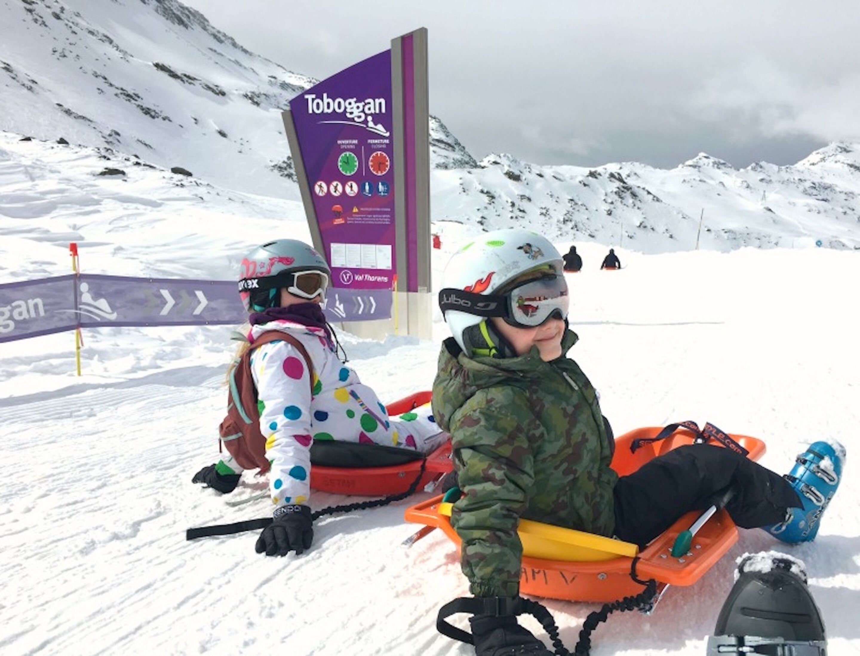 Deux personnes dévalent une pente enneigée sur une luge, avec un panneau indiquant « Toboggan » en arrière-plan.