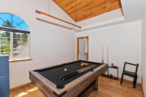 Una mesa de billar con bolas y tacos, una ventana con vistas a la vegetación, y una puerta en una habitación con suelo de madera.