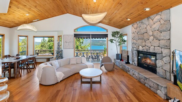 A spacious living area with a stone fireplace, wooden ceiling, and large windows offering a view of the outdoors.