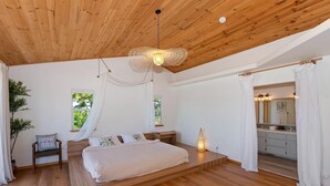 A bedroom with a wooden ceiling, a bed, a bedside table, a chair, a window, and a bathroom with a mirror.