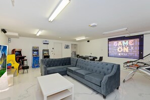 Una habitación con sofá modular, juegos de arcade, una pantalla grande mostrando 'GAME ON' y una mesa de centro blanca.