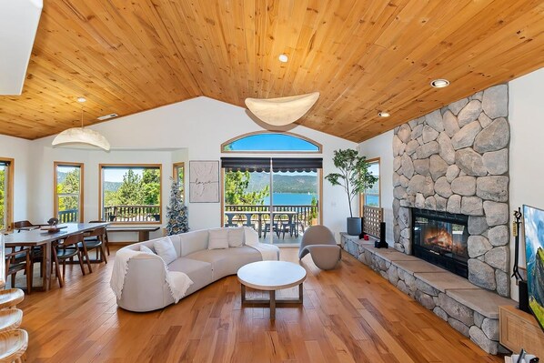 A spacious living room with a stone fireplace, wooden ceiling, and large windows.