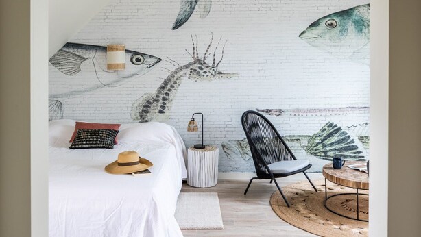 Ein Schlafzimmer mit schrägem Dach, einem Bett mit weißer Bettwäsche, einem Stuhl, einem kleinen runden Tisch und einem Wandgemälde mit Fischen.