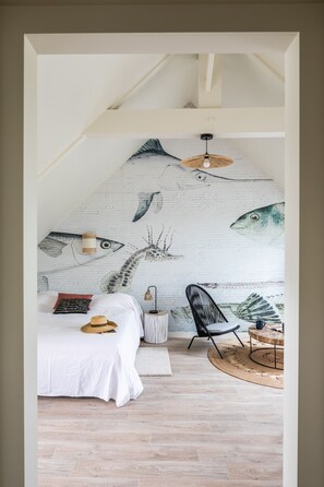 Ein Schlafzimmer mit schrägem Dach, einem Bett mit weißer Bettwäsche, einem Stuhl, einem kleinen runden Tisch und einem Wandgemälde mit Fischen.