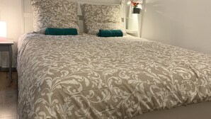 A neatly made bed with a patterned bedspread and two teal pillows, a bedside table with a lamp, and a wall-mounted nightstand with a small plant. - Historical Town Center Cosy Flat (Colmar)