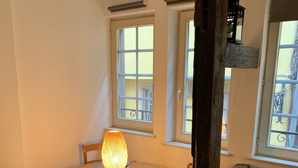 A cozy dining area with a round wooden table, four chairs, and a lit table lamp. There are two windows with white frames, one of which has a view of balconies. A wooden beam and a hanging lantern are visible. - Historical Town Center Cosy Flat (Colmar)
