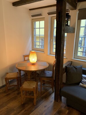 A cozy dining area with a round wooden table, four chairs, and a lit table lamp. There are two windows with white frames, one of which has a view of balconies. A wooden beam and a hanging lantern are visible.