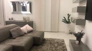 A modern living room with a sectional sofa, a flat-screen TV mounted on the wall, a small side table with decorative items, and a potted plant. - La Casa dei Gemelli (Priolo Gargallo)