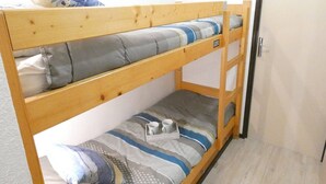 Een stapelbed met houten frame, beddengoed in grijs en blauw, en een klein tafeltje met een kopje en een doos tissues.
