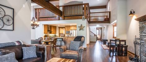 A spacious living area with a high ceiling, wooden beams, a staircase, a fireplace, and a kitchen in the background.