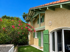 A building with a green door, a window with a white frame, and a small garden area with green grass and pink flowers. - Spacious villa with pool for 8 pers in Soustons (Soustons)