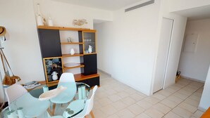 A dining area with a glass table and white chairs, a wooden bookshelf with decorative items, and a white tiled floor. - Superb air-conditioned apartment with large terrace and parking in Menton (Menton)