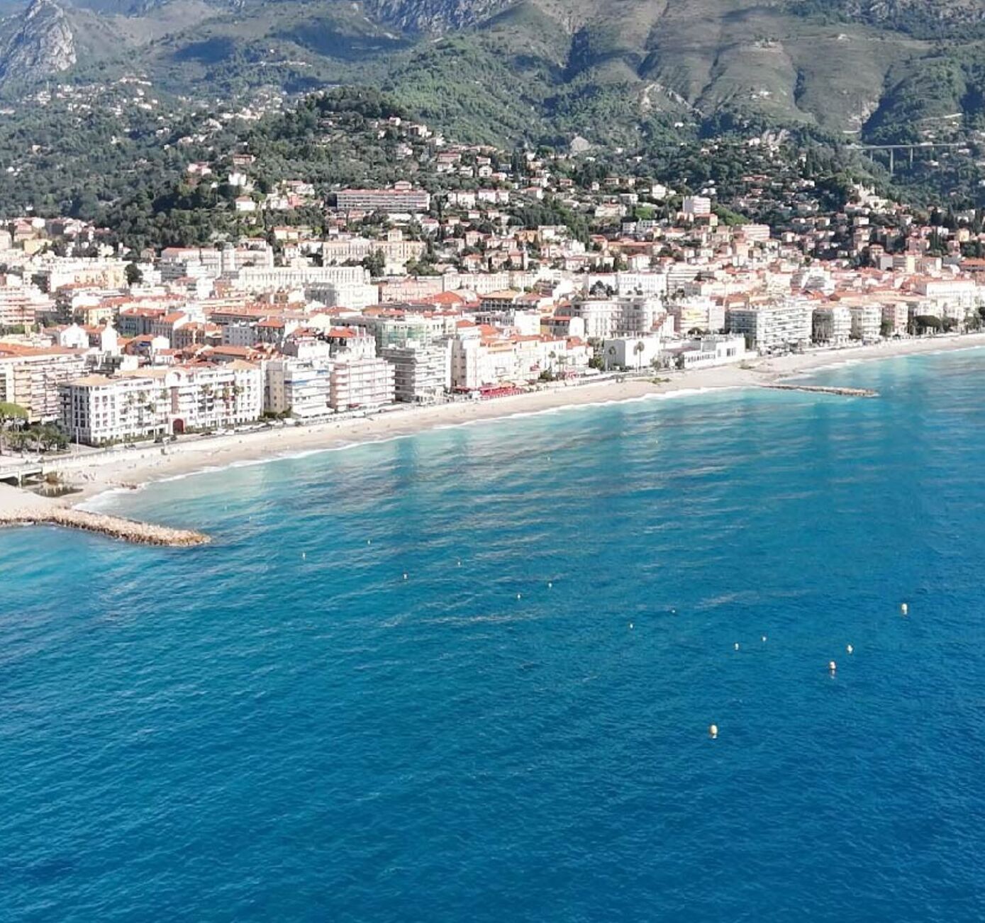 Eine Küstenstadt mit Gebäuden entlang der Uferlinie, blauem Meer und Bergen im Hintergrund.