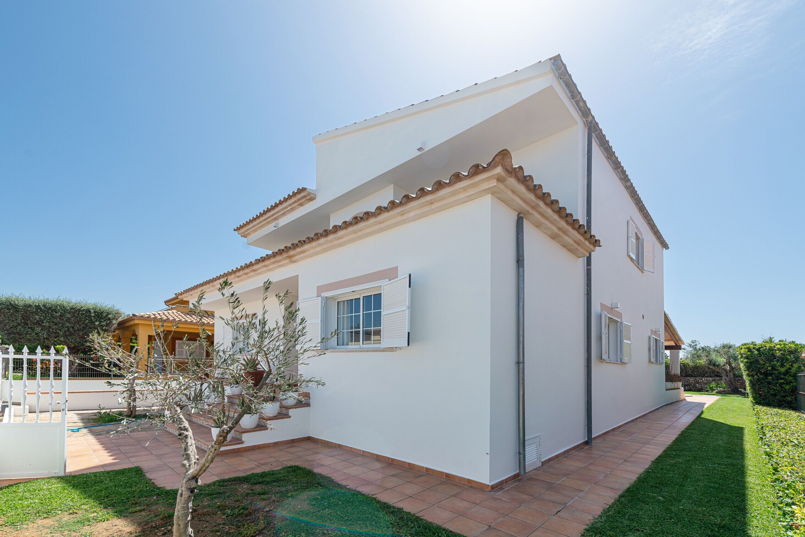A two-story house with a white exterior, a tiled patio, and a small tree in the front yard.