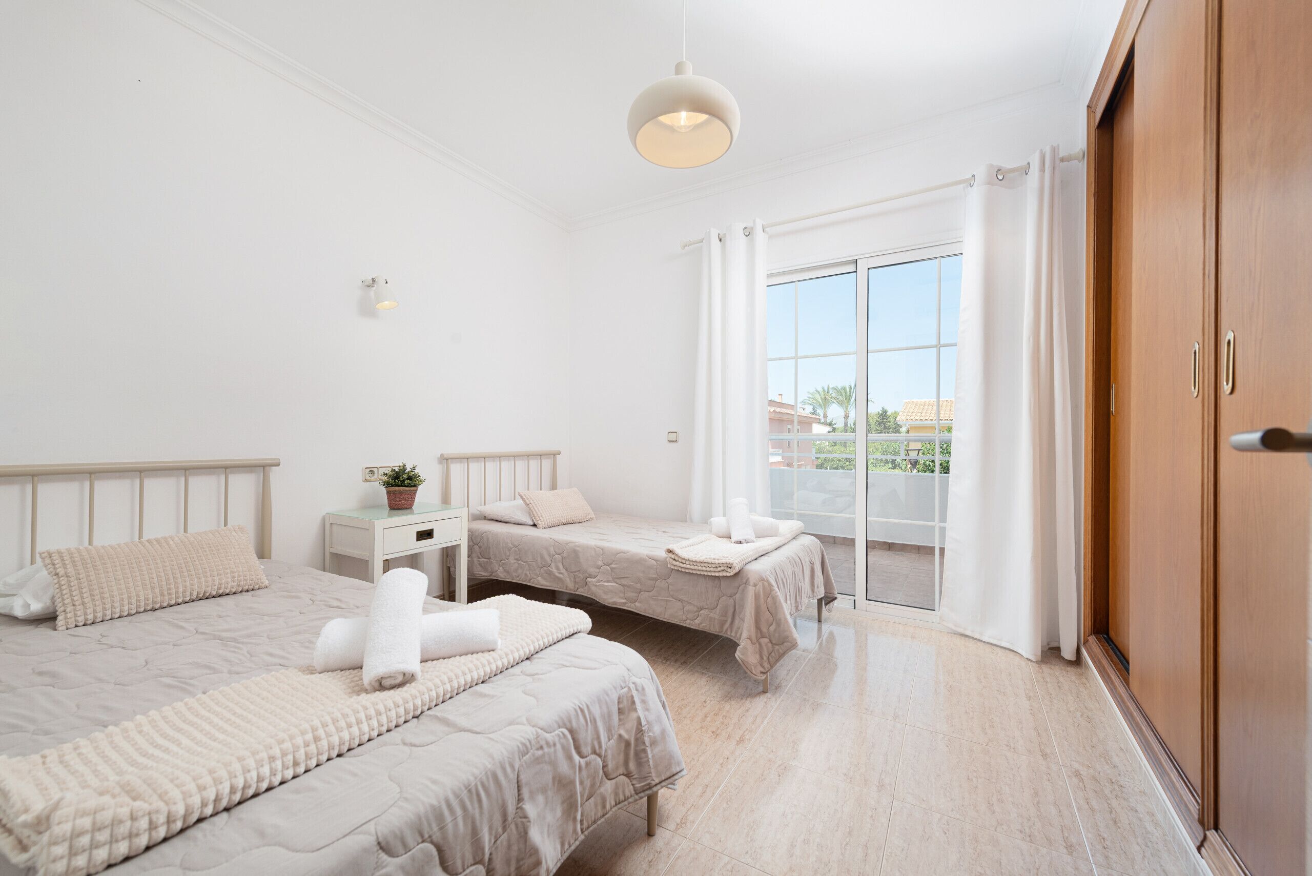 A bedroom with two beds, a bedside table with a plant, and a large window with curtains.