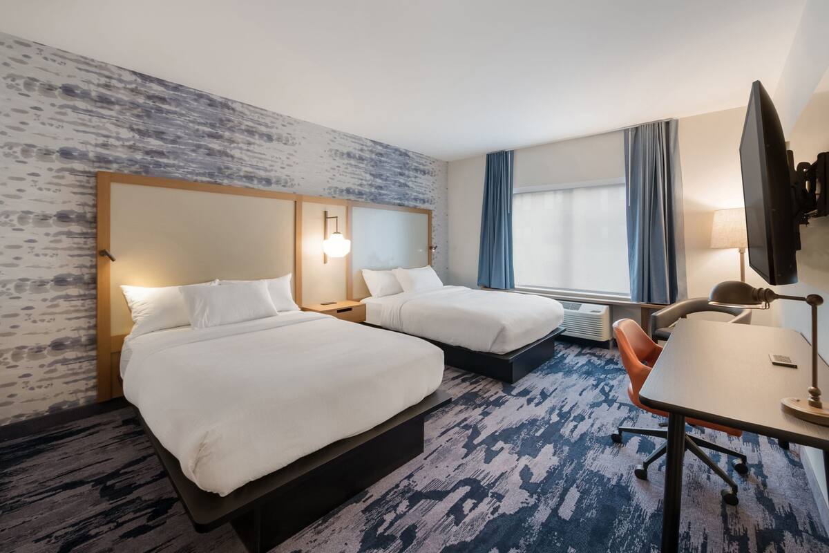 Une chambre d’hôtel avec deux lits, un bureau, une chaise, une télévision et une grande fenêtre avec des rideaux.