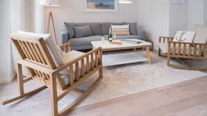 Una sala de estar moderna con un sofá gris, sillas mecedoras de madera, una mesa de centro blanca y un cuadro con una vista urbana en la pared.