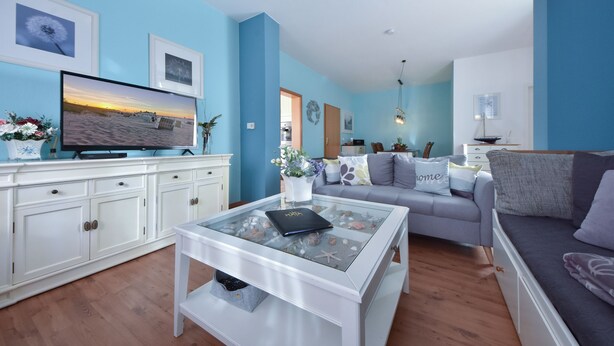 A modern living room with a flat-screen TV, a glass coffee table, a sofa, and a dining area with a white cabinet and a vase of flowers. - Villa zur See 10 - modern maritime vacation apartment (Heringsdorf (Seebad))
