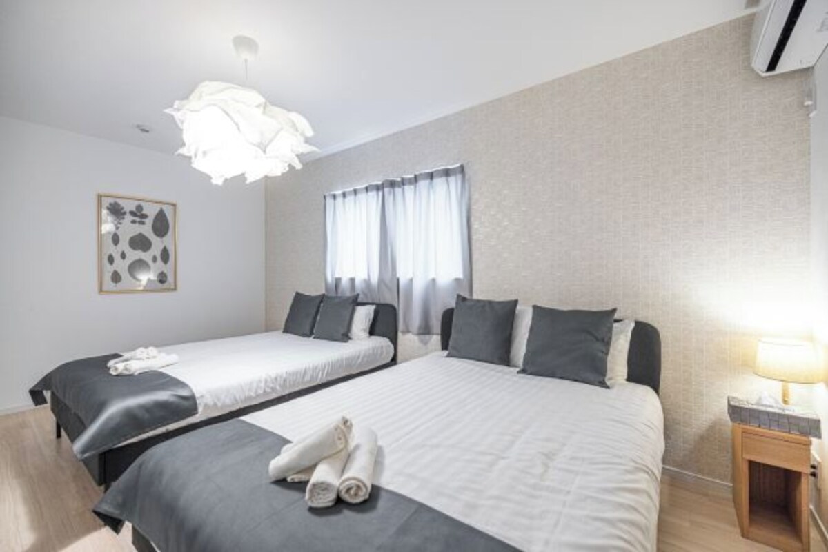 Ein Hotelzimmer mit zwei Betten, einem Nachttisch mit Lampe, einem Fenster mit Vorhängen und einer wandmontierten Klimaanlage.
