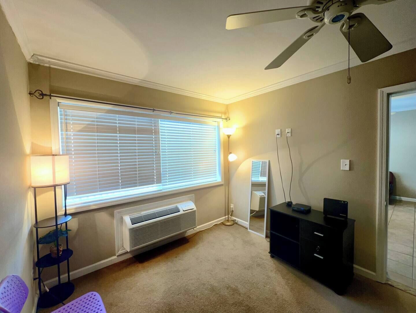 Una habitación con ventilador de techo, un ventanal con persianas, un calefactor portátil, un escritorio con computadora y un aparador negro.