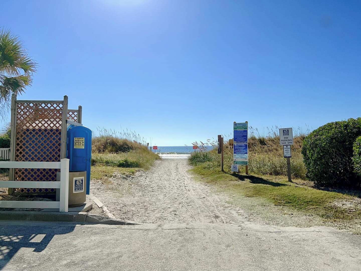 Un sentier sablonneux mène à une plage bordée d’une clôture en bois et, sur la gauche, on aperçoit une boîte de couleur bleue, probablement un coffret de raccordement électrique.