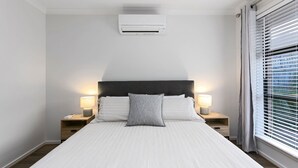 A hotel room with a bed, two bedside lamps, a window with blinds, and an air conditioning unit.
