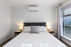 A hotel room with a bed, two bedside lamps, a window with blinds, and an air conditioning unit.