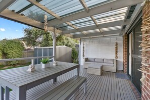A covered outdoor area with a wooden deck, a seating area with cushions, and a wall with hanging decorations.
