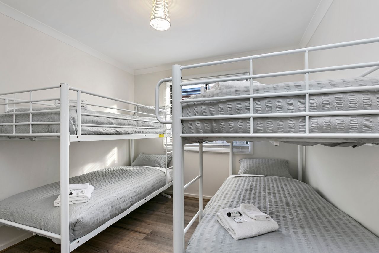 Ein Schlafraum mit Etagenbett, einem weißen Metallgestell, grauer Bettwäsche und einem kleinen Nachttisch mit Lampe.