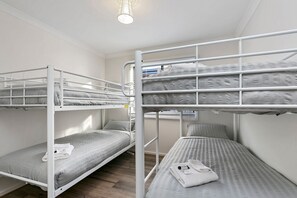 A bunk bed room with a white metal frame, grey bedding, and a small bedside table with a lamp.