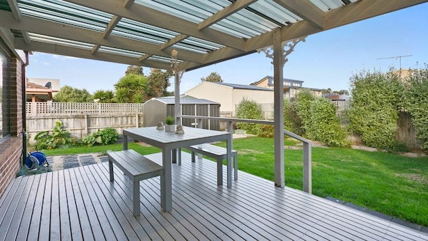 A covered outdoor deck with a table and benches, surrounded by a grassy yard and a wooden fence.