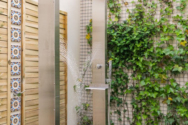A shower with a vertical water stream, a wooden door with decorative tiles, and a wall covered with green ivy.