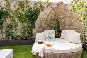 A wicker daybed with a canopy, surrounded by lush green ivy, and a white hat and book on the bed.