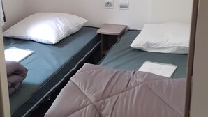 A dormitory room with three single beds, each with a pillow and a blanket. There is a small table between the beds, and a shelf above the beds.
