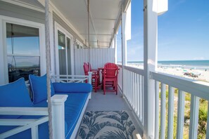 A covered porch with a view of the beach, a white railing, and a red and blue outdoor seating area. - Spacious Oceanfront and Pier Views~ Seaside Villas 202 (Folly Beach)