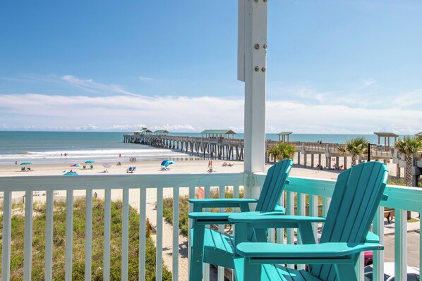 A beach view with a pier extending into the ocean, turquoise chairs, and a white fence. - Spacious Oceanfront and Pier Views~ Seaside Villas 202 (Folly Beach)