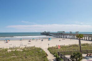 A beach with a wooden pier extending into the ocean, surrounded by beach chairs and umbrellas. - Spacious Oceanfront and Pier Views~ Seaside Villas 202 (Folly Beach)