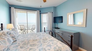 A bedroom with a bed, a TV, a dresser, and a balcony with chairs. - Spacious Oceanfront and Pier Views~ Seaside Villas 202 (Folly Beach)